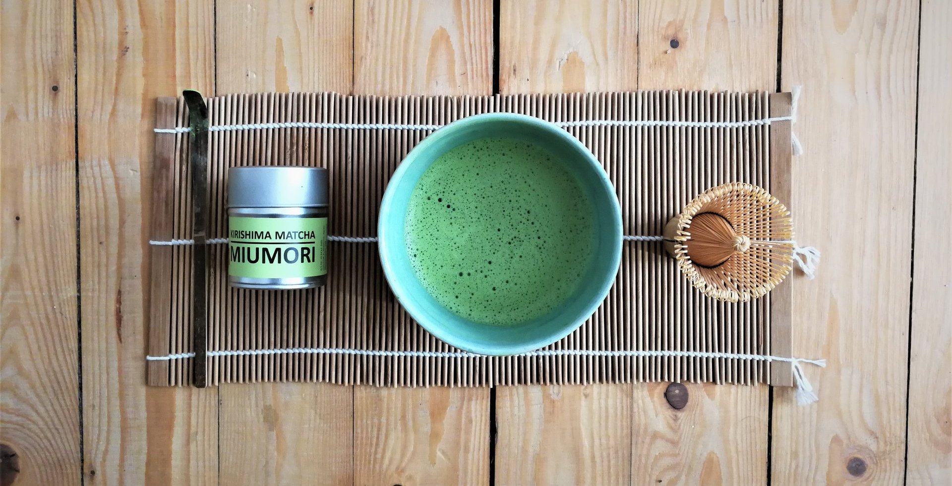 Matcha-Tee-Zubereitung mit Zubehör: eine Dose Kirishima Matcha Miumori, ein traditioneller Bambusbesen (Chasen), ein Bambuslöffel (Chashaku) und eine Schale mit aufgeschäumtem, grünem Matcha-Tee – alles auf einer Bambusmatte arrangiert