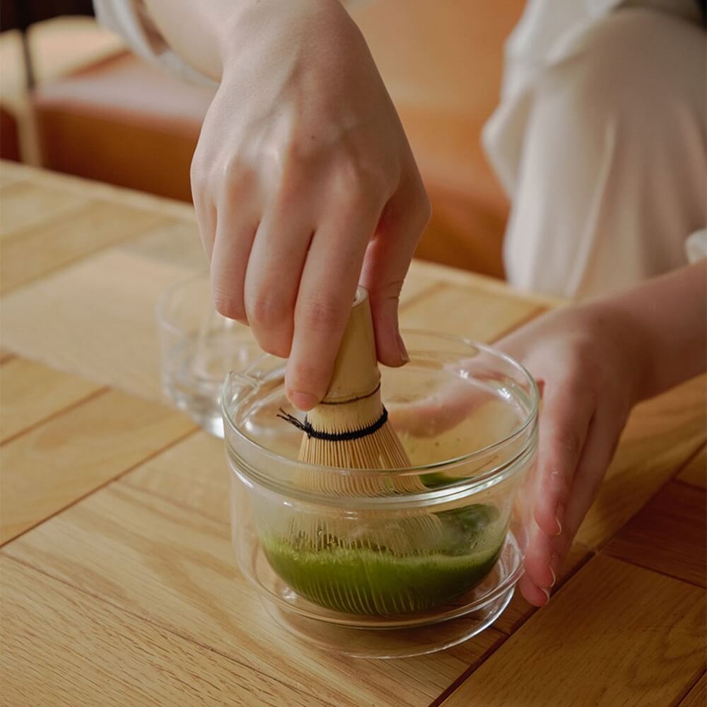 Matcha wird mit Bambusbesen in einer doppelwandigen Matchaschale aus Glas aufgeschlagen
